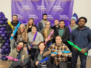 Part of the Credico team poses as a group in front of a Cradles to Crayons backdrop while smiling and holding large plastic crayons.