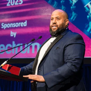 Obiora Shorinwa, wearing a navy tuxedo with black lapels, gives a speech on stage behind a podium following his recognition as a Regional Consultant at the January 2023 North American Awards Gala in Miami, Florida