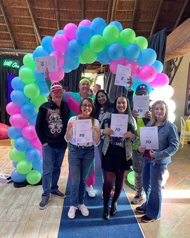 Credico South Africa staff recognized for milestone years of service pose for a group photo while holding their certificates under a brightly-colored balloon arch at the company's annual Christmas in July celebration in 2024.