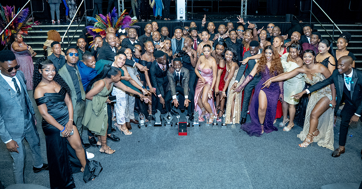 A large group of independent business owners in glamourously sparkling gala attire gather for a group photo in a semicircle with all their awards in a row in front of them, pointing to emphasize their success and recognition