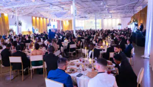 A wide view of the room full of independent business owners, friends, guests, and supporters seated around round, gold-adorned tables during the 2026 Limitless Kickoff Gala. Credico founder and chairman Antoine Nohra can be seen on stage to the left, in front of a large video screen surrounded by golden curtains.