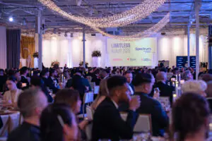 A wide-angle view of the crowd of independent business owners in their formal attire seated for dinner at the 2026 Limitless Kickoff Gala. White draperies and twinkle lights adorn the walls and ceiling, and a gold-branded Limitless Kickoff sponsorship banner is displayed on the back wall.