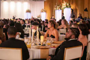Attendees watch raptly as top talent is recognized at the 2026 Limitless Kickoff Gala. Everyone is in formal attire and seated around large round tables with gold place settings and towering candles, and lighted pedestals topped with massive floral arrangements line the back of the room.